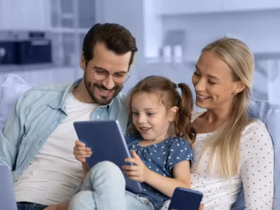 father, mother and young girl sitting on a sofa looking at an iPad the child is holding.