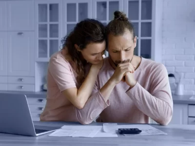 Unhappy young woman supporting worried husband looking at documents on a kitchen counter.